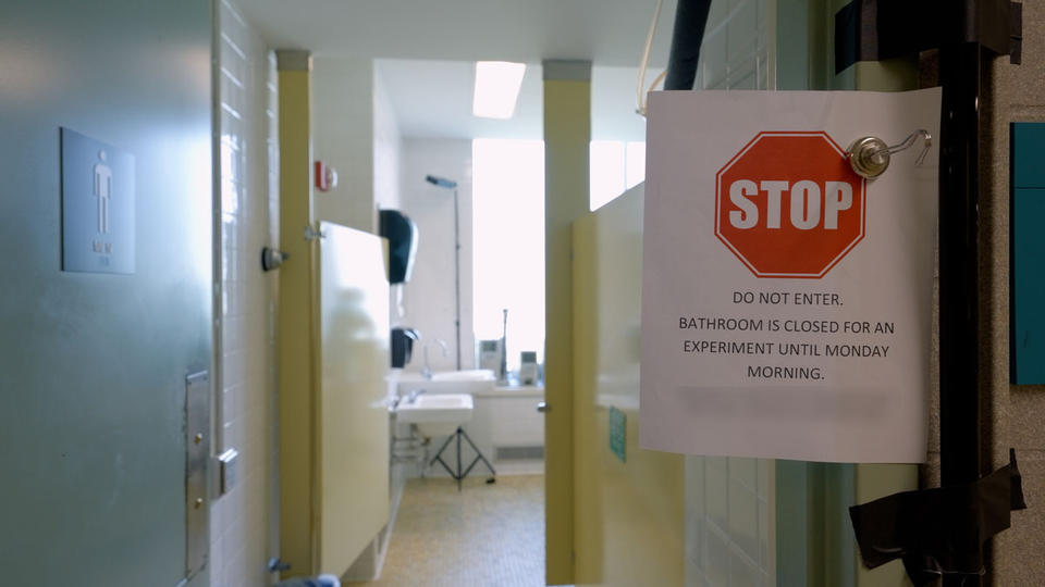 A paper sign is clipped in front of a open door to a public restroom. The sign reads, “Stop. Do Not Enter. Bathroom is closed for an experiment until Monday Morning.”