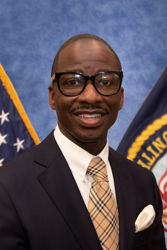 A picture of an African American male wearing black-framed eyeglasses, a blue suit jacket, a white collared shirt, a brown necktie, and a white pocket square.