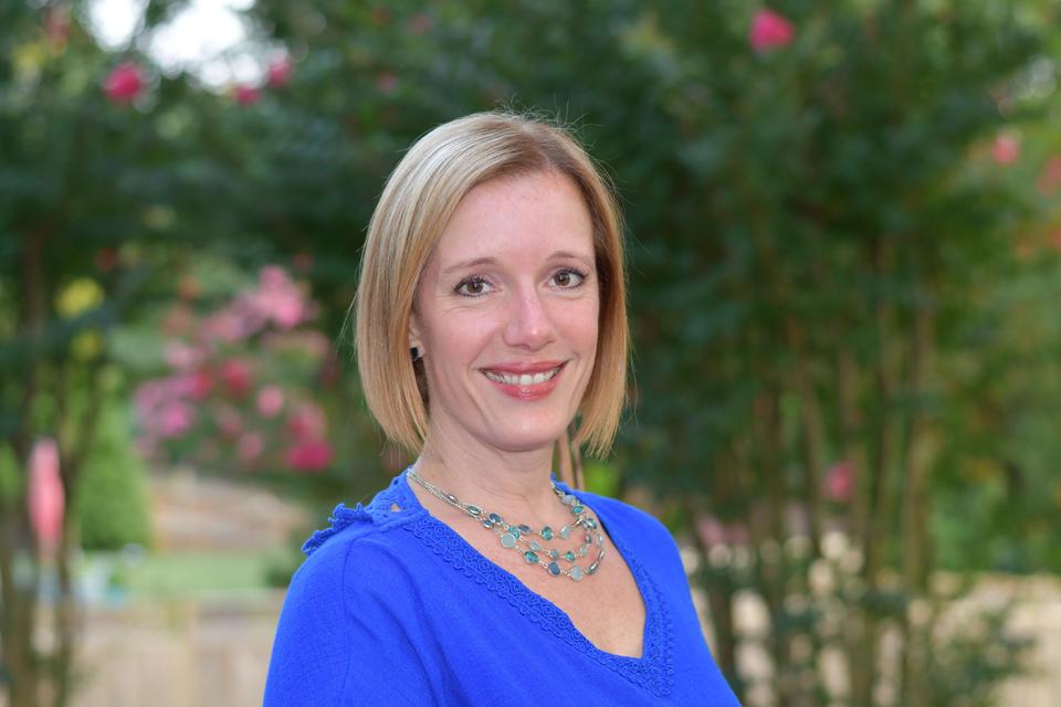 smiling woman with short blonde hair wearing a bright blue shirt with trees and pink flowers in the background