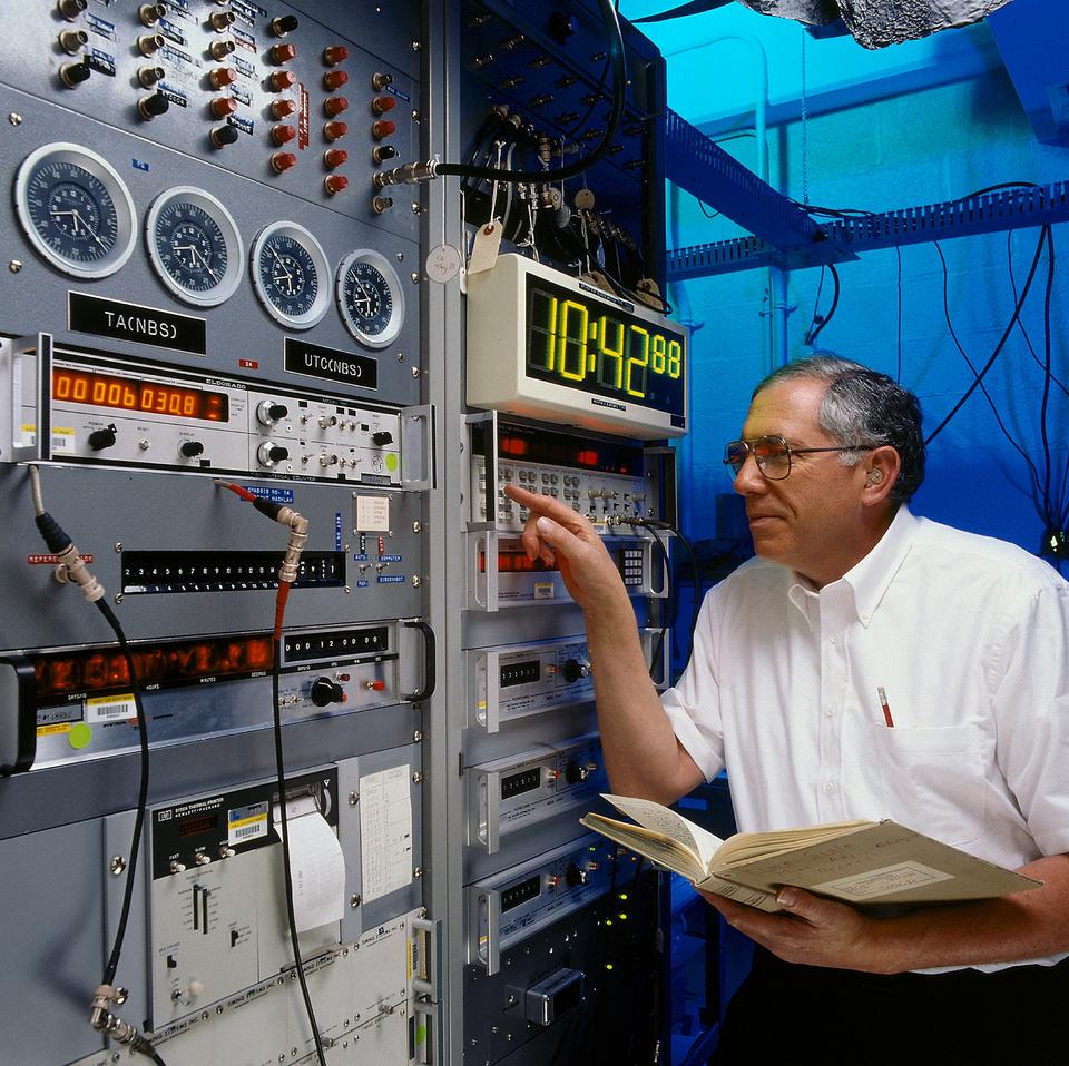 A researcher holding an open book points at a button on a large, complex, retro-looking machine featuring a digital clock readout. 