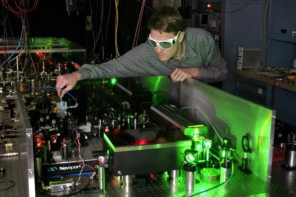 A researcher wearing safety glasses leans over a table crowded with laser devices in a darkened lab. 