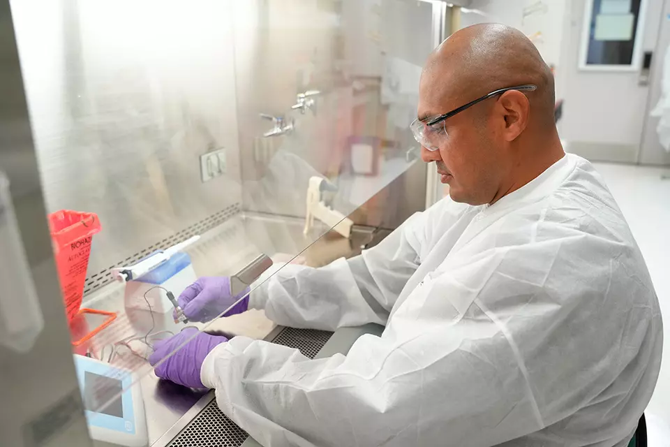 A researcher wearing safety gear works with small tools in the lab, putting his hands inside a plexiglass box. 
