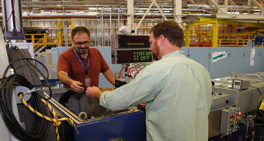 Two men working on opposite sites of the nSoft neutron beam station. In the background there are lots of pipes, shelving, etc. Gives the appearance of a warehouse type setting.