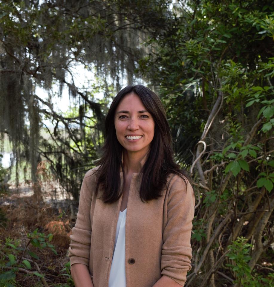 A woman with shoulder length black hair wearing a tan blazer and white shirt standing in front of a marshland.