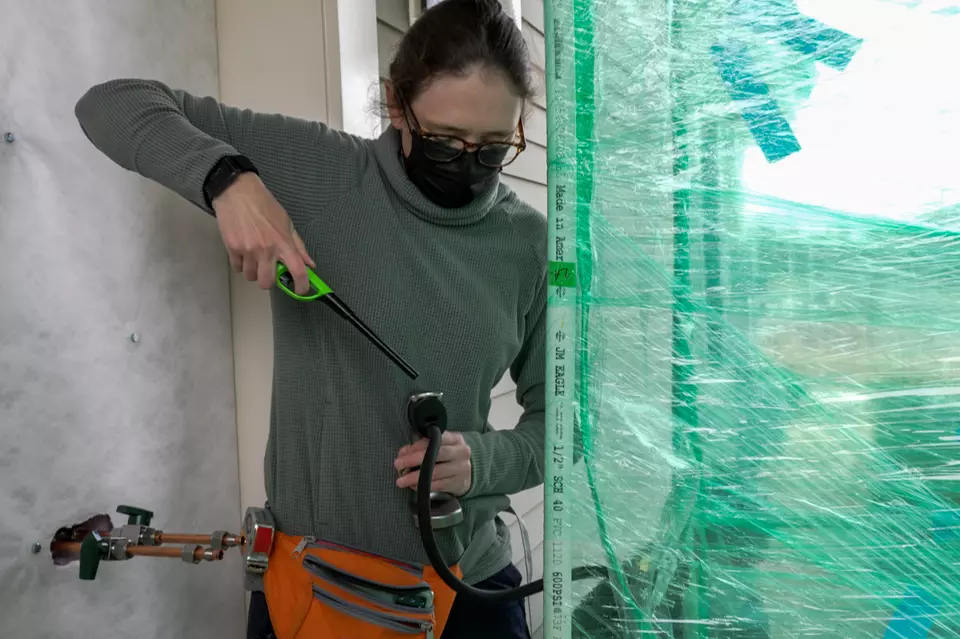 A researcher standing outside a house holds a lighter to the end of a hose-like device leading into an area closed off by plastic wrap. 