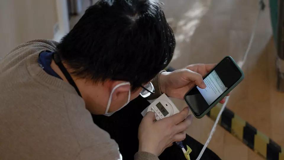 A researcher bends over a digital readout on a piece of equipment, comparing it to information on his phone. 