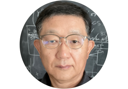 person with short black hair and wearing glasses, standing in front of a chalk board covered in calculations