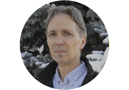 person with short grey hair wearing a dress shirt, standing in front of snow-covered evergreens