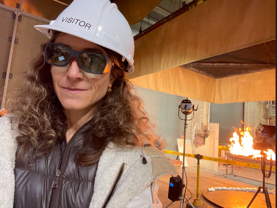 Esther Baumann wears a white helmet and safety goggles as she poses in front of a flaming experiment in the fire lab. 