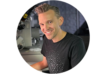 Andrew D. Iams, a person with short dirty blonde hair, sitting at a computer and smiling