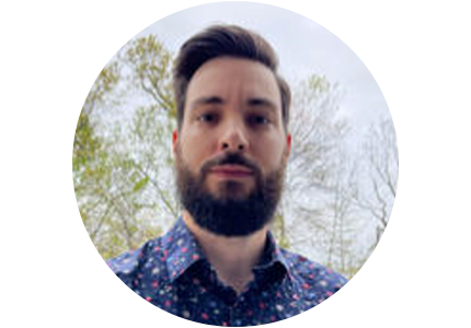 person with dark brown hair and a beard, wearing a dress shirt and standing in front of trees