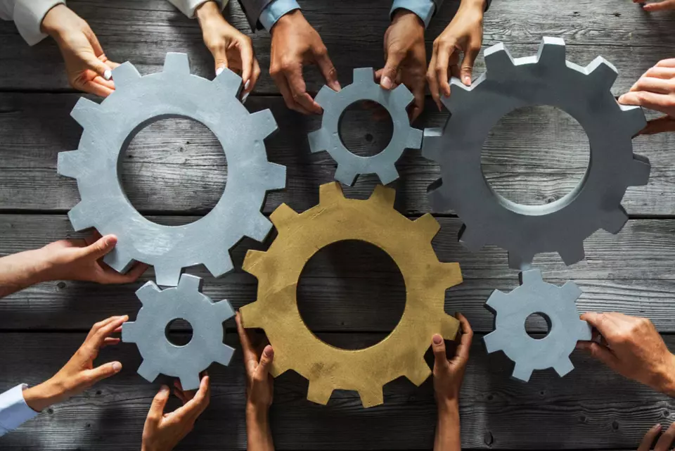 people gathered around a table push large gears together, to symbolize teamwork and collaboration