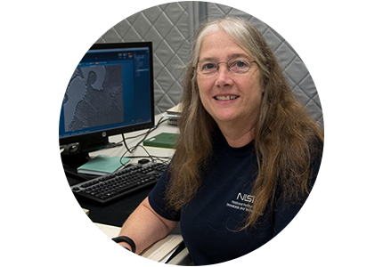 NIST's Maureen Williams, a person with long brown/gray hair and glasses, sits at a computer with technical graphics and smiles for the camera