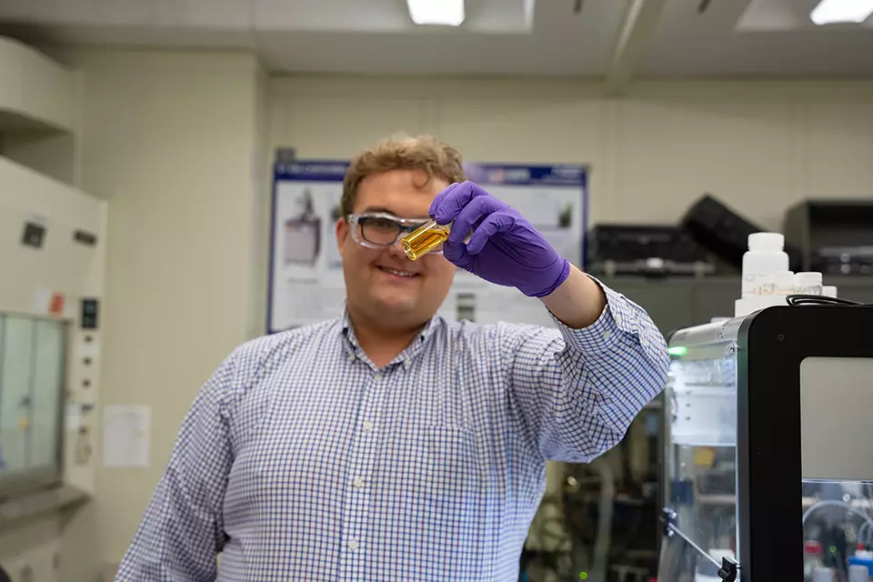 Peter Beaucage stands in the lab holding up a vial of yellowish liquid. 