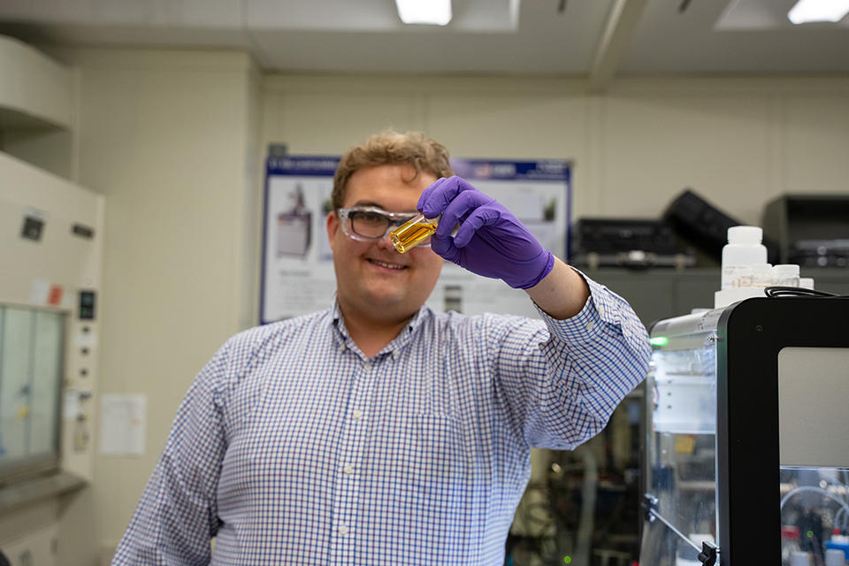 Peter Beaucage stands in the lab holding up a vial of yellowish liquid. 