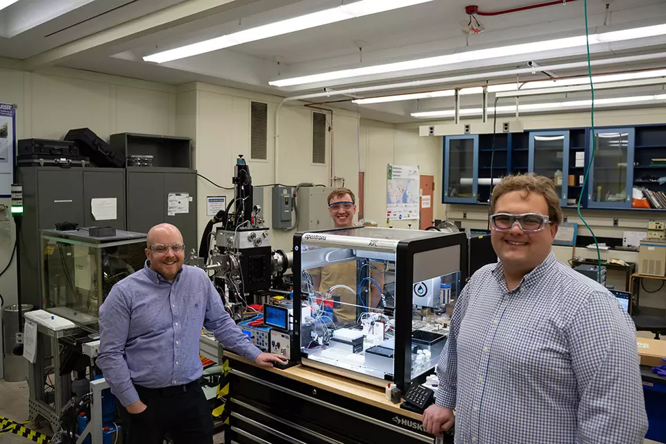 Three men in safety glasses pose standing around a large clear plastic box on a lab table. The box holds several small pieces of equipment. 