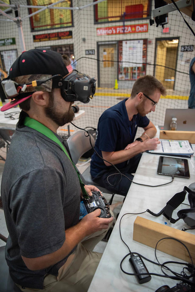 A man wearing an electronic device over his eyes sits at a table holding a controller for a drone. The indoor training facility includes pretend storefronts like a pizza shop. 