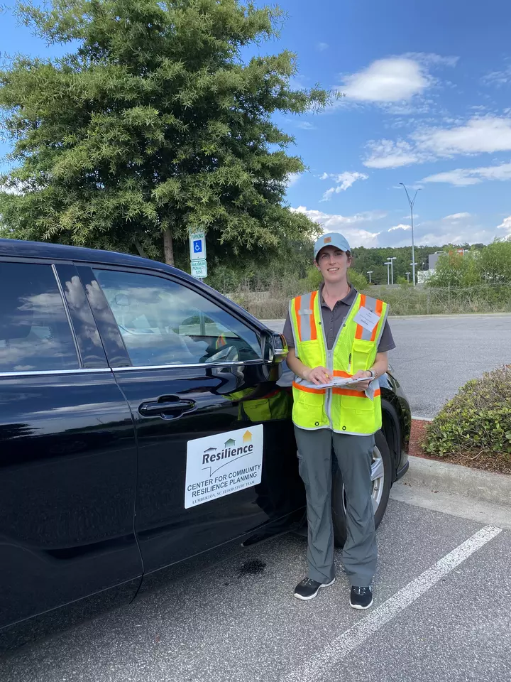 Jennifer Helgeson, holding a clipboard and wearing a high-vis vest stands next to a car. The car has a large sign on the side that reads “Center for Community Resilience Planning. Lumberton, NC Flood Study Team.” 
