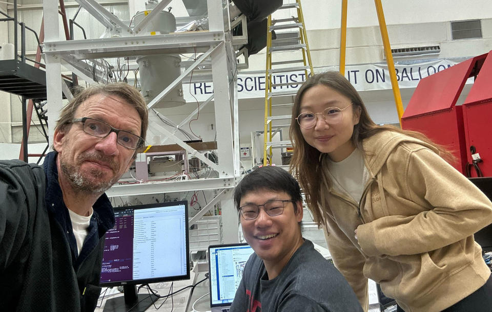Three researchers lean into the frame for a group shot, set inside a large lab space with ladders in the background.