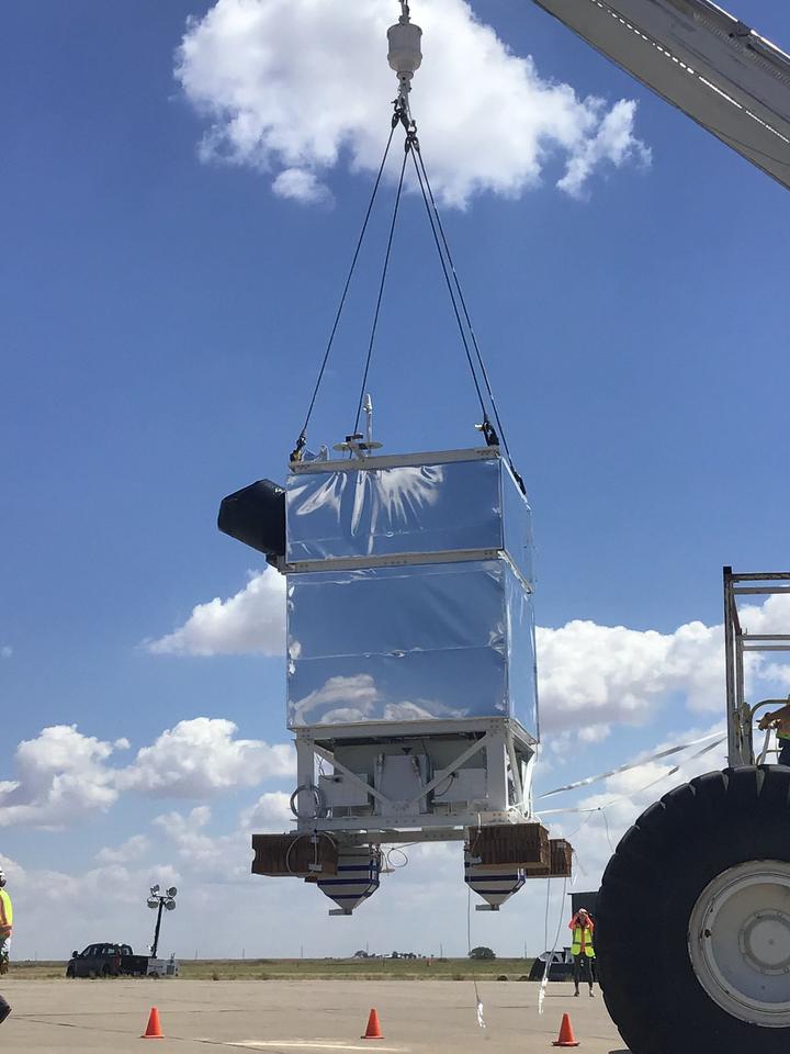 A rectangular box coated with reflective foil hangs from cables over a parking lot.