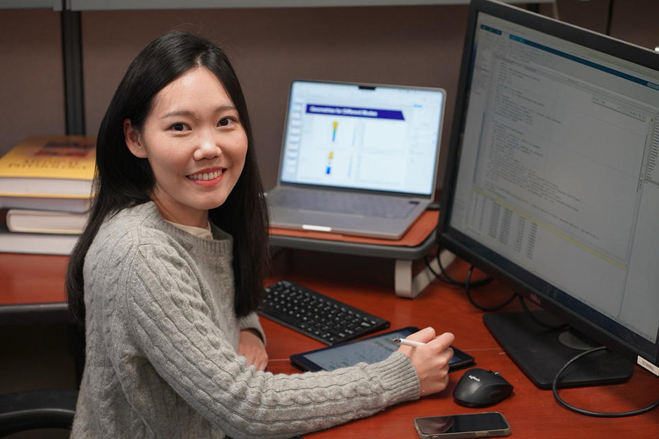 A woman poses smiling, sitting at a desk with a laptop and an additional large monitor. 