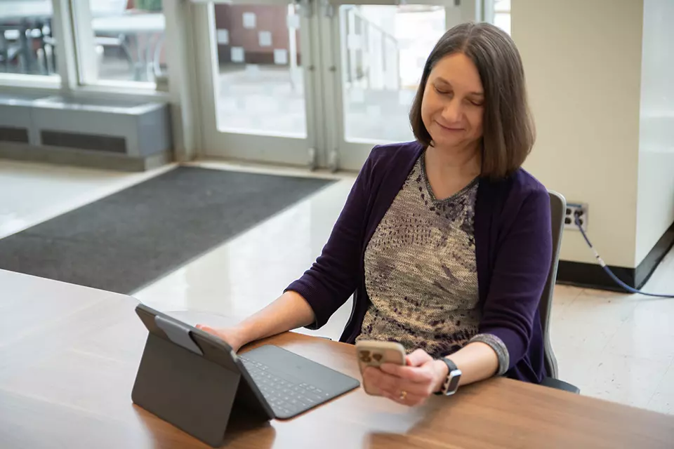 Julie Haney sits at a table in the NIST library with a laptop, looking at a smartphone in her hand. 