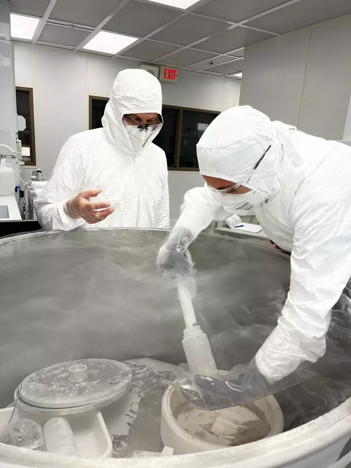 Researchers in white hazmat coveralls reach into an open freezer with a scooping device as vapor rises from the freezer.