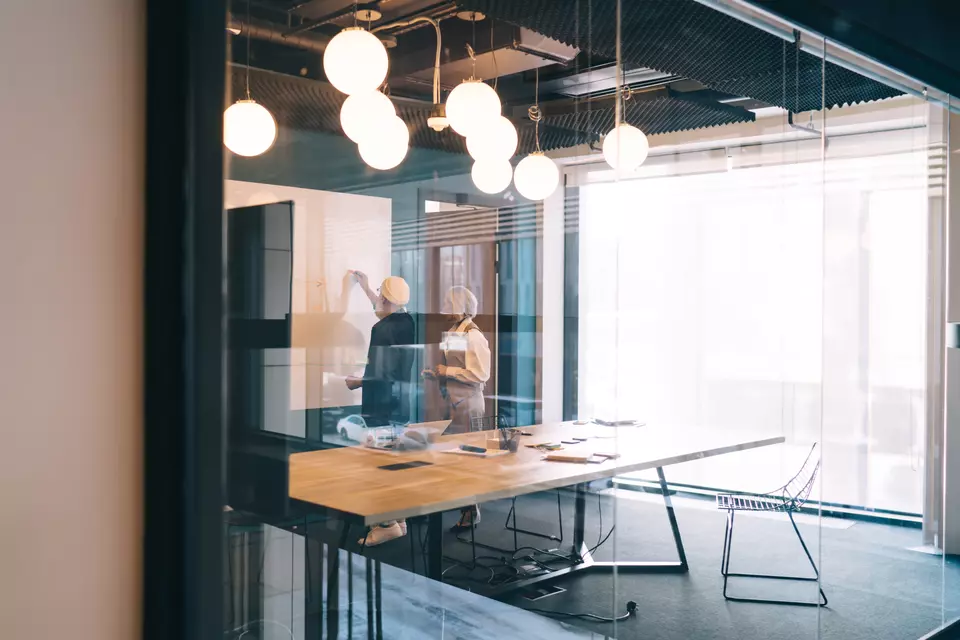 At the far end of a modern conference room with overhead globe lighting, two people stand looking at a whiteboard. 