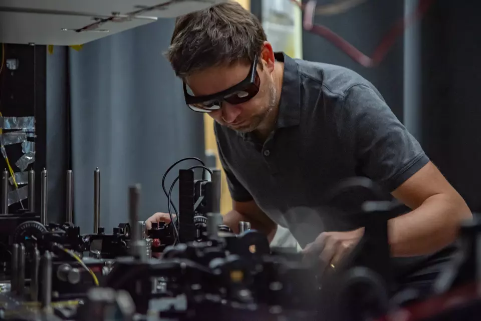 Krister Shalm wears safety glasses as he leans over a lab table covered with electronics equipment.