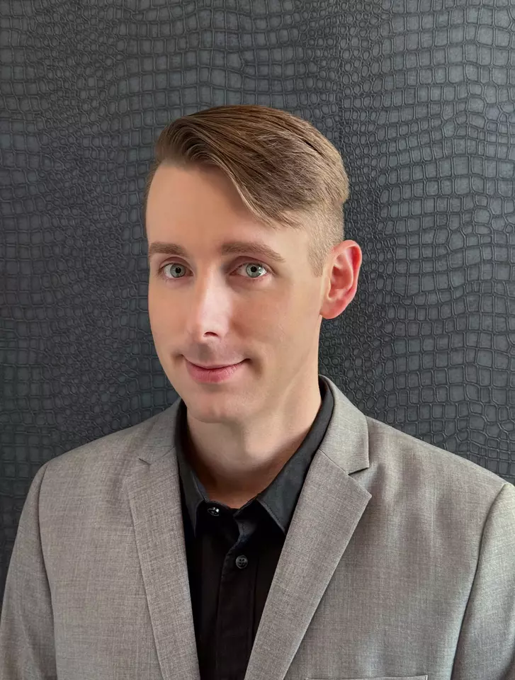 Headshot of Kyle Anderson, a person with short blonde comb over hair, wearing a black collared shirt and a gray suit jacket, smiling