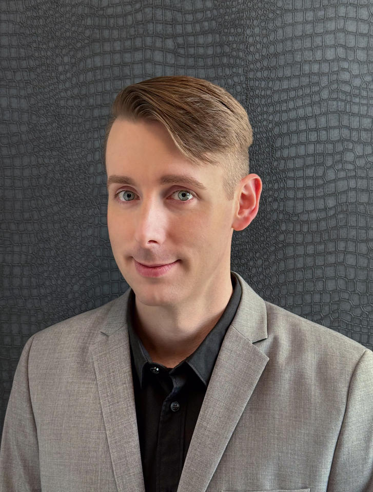 Headshot of Kyle Anderson, a person with short blonde comb over hair, wearing a black collared shirt and a gray suit jacket, smiling