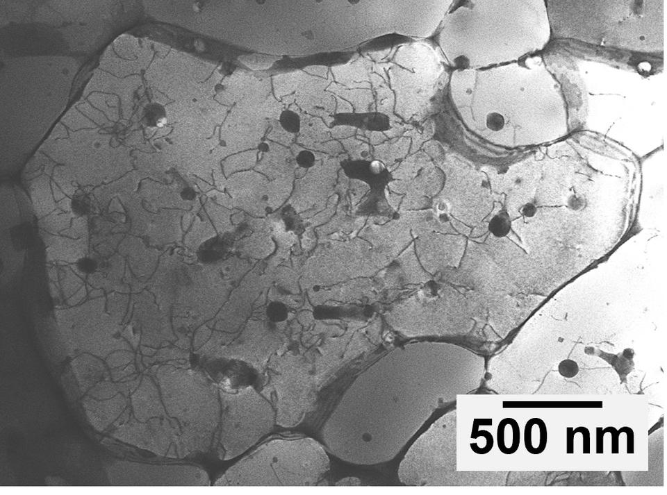A black and white image of the aluminum alloy under a microscope. Most of the image is light grey, but there are many small black spots that have lines extending out from them like cracks.