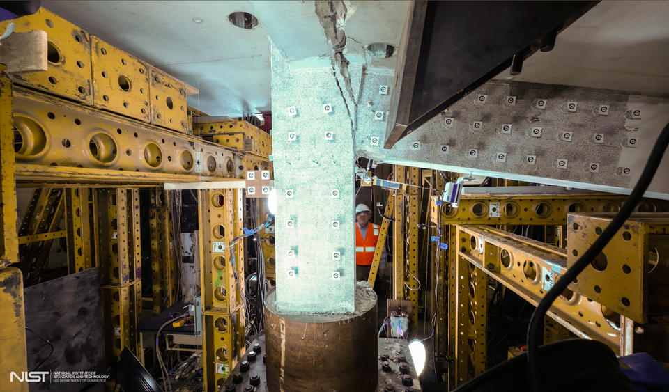 Heavy equipment and scaffolding surrounds a vertical concrete slab and attached horizontal beam, which is being cracked away.