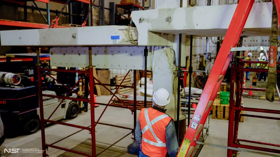 A person wearing construction safety gear looks up at a cracked concrete slab and beam arrangement held up by scaffolding in a large indoor space. 