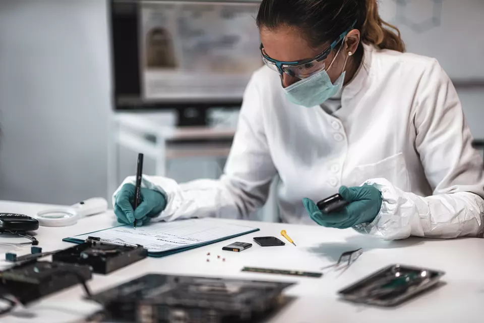 A researcher wearing a mask, safety glasses and gloves sits at a desk, looking at electronic devices and writing on a clipboard. 