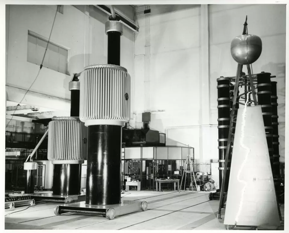 Historical photo shows a large lab space with cylindrical metal towers of different shapes. 