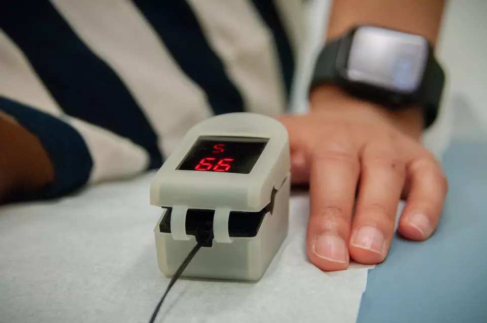 Close-up of a clip-on medical device on the finger of a person sitting on an exam table. The device screen reads 99. 