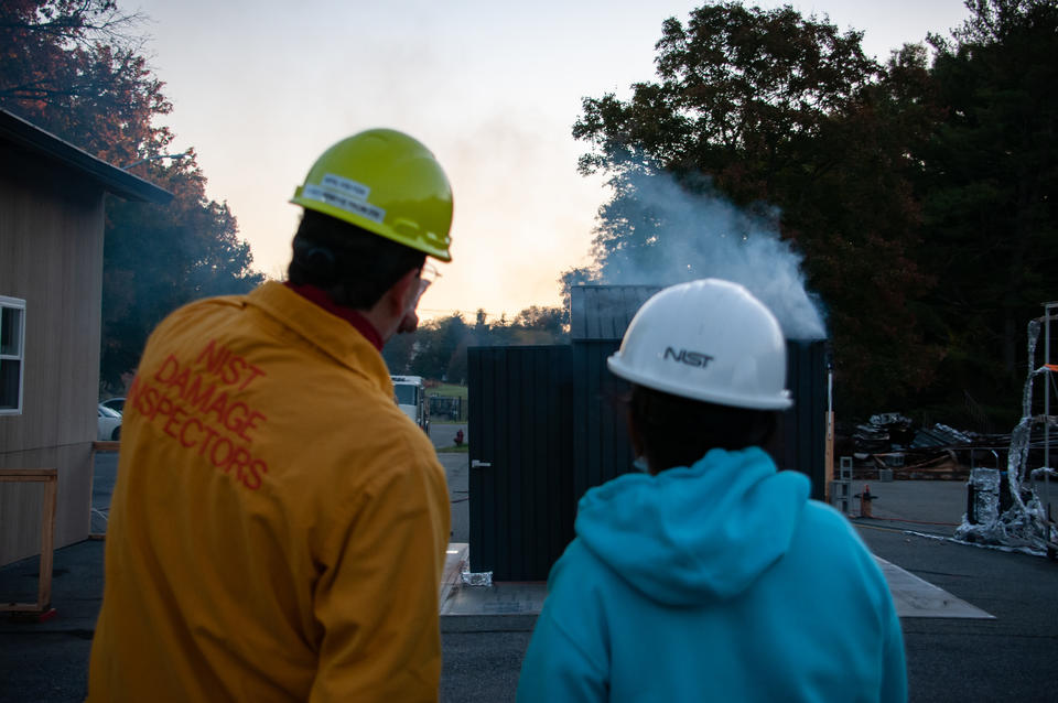 Two NIST researchers, wearing hardhats and protective jackets, watch a small shed burn as part of a setudy.