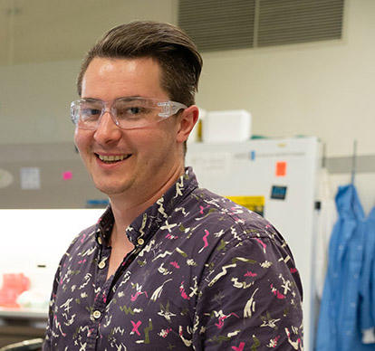 Sam Schaffter wears safety glasses as he poses smiling in the lab.