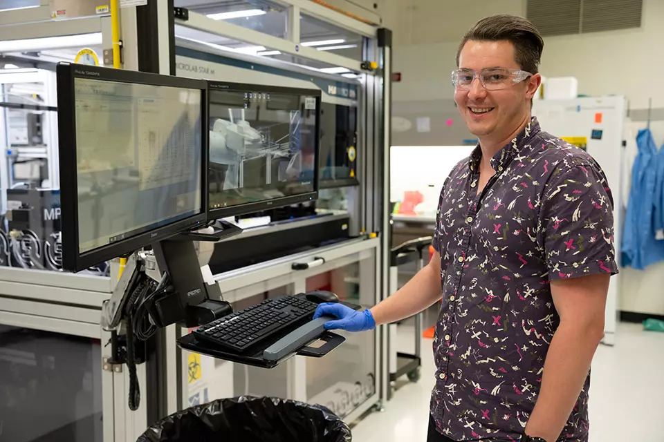 Researcher wearing safety glasses stands next to a computer in the lab, with a large glass-walled device in the background.