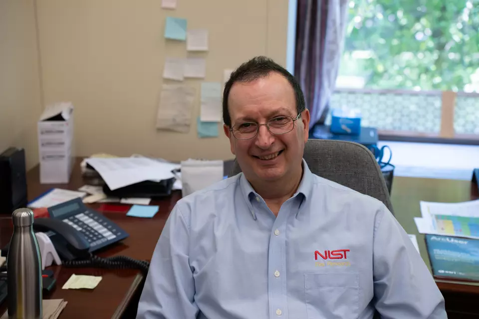 A man wearing a NIST collared shirt sits in an office and looks at the camera