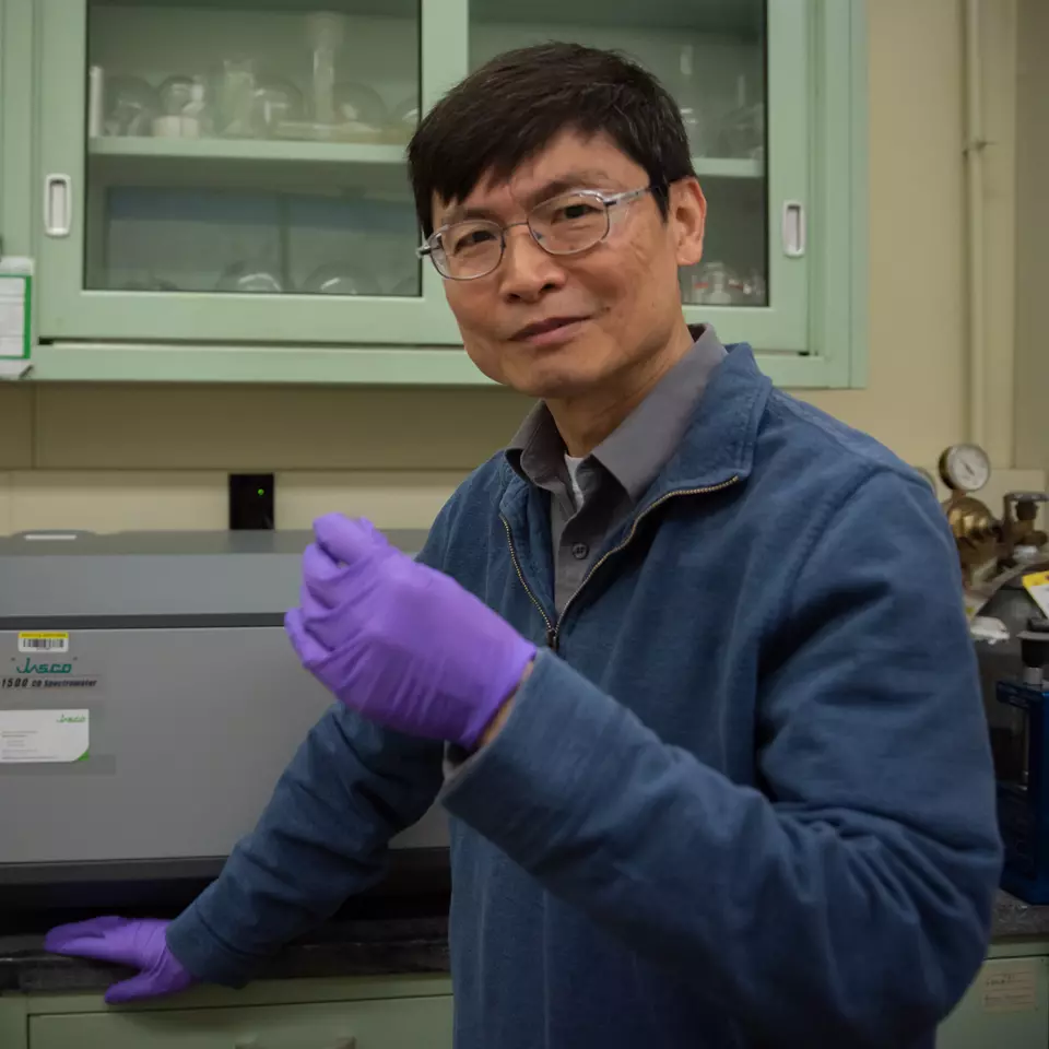 Researcher Ming Zheng looks at the camera as he stands in the lab holding a sample in his gloved hand. 