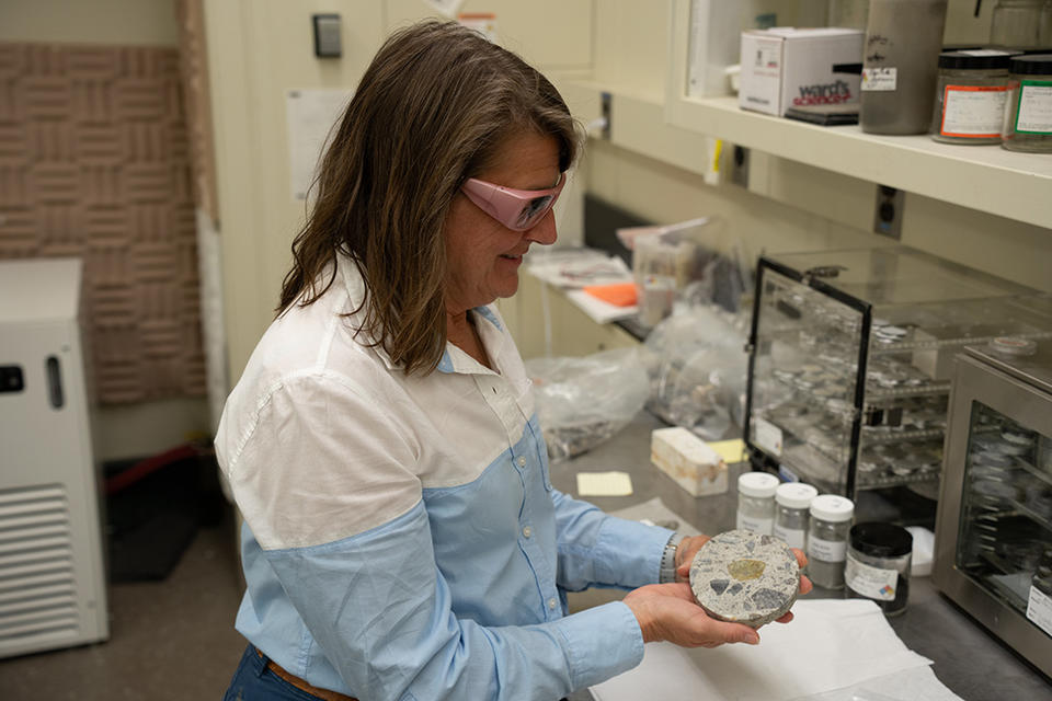 A researcher wearing safety glasses stands in the lab displaying a small disk of concrete with visible rocks inside it. 