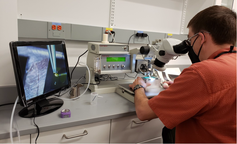 A researcher peers into the eyepiece of a microscope while positioning a small box beneath it; the image shows on a monitor to his left.
