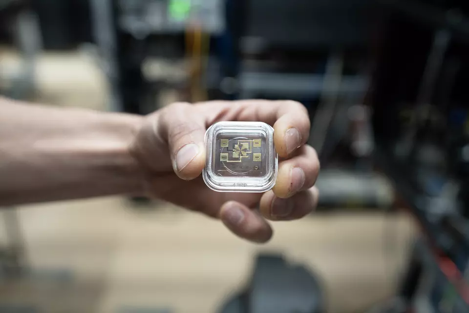 A hand holds a small square device in a clear case. Gold-colored electronics can be seen through the case. 
