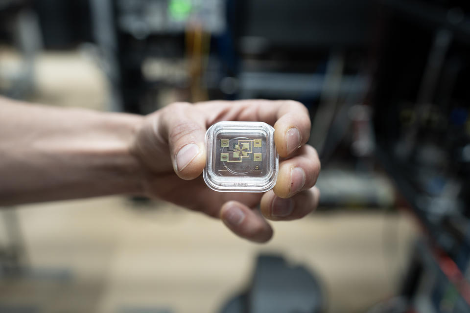 A hand holds a small square device in a clear case. Gold-colored electronics can be seen through the case. 