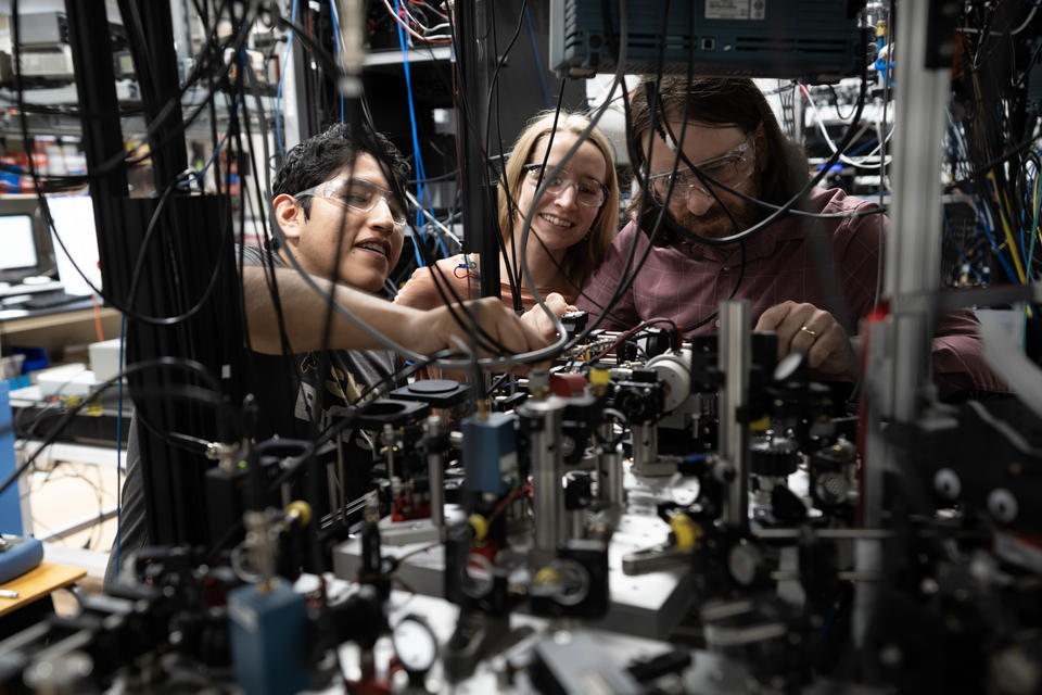 Three researchers in safety glasses are seen through a forest of hanging wires and other electronic and laser devices. 