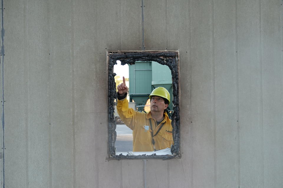 A man wearing a yellow hardhat and fire-resistant jacket points to burnt frame of a window.