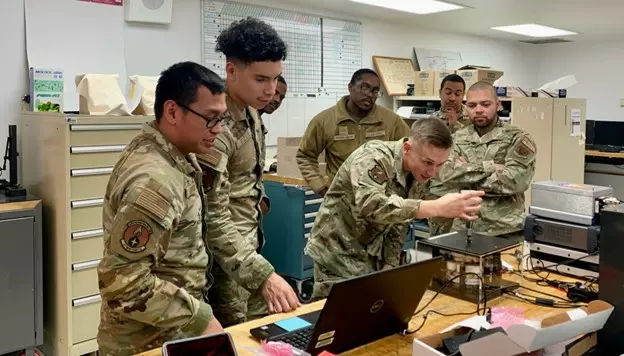 Airmen in uniform gather around a desk that holds an open laptop and several electrical devices, including one inside a clear box. 