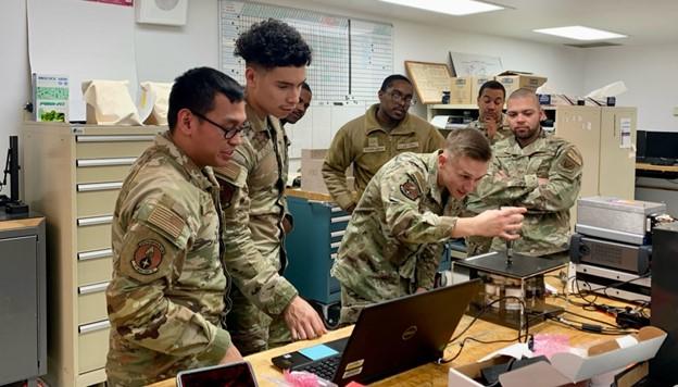 Airmen in uniform gather around a desk that holds an open laptop and several electrical devices, including one inside a clear box. 